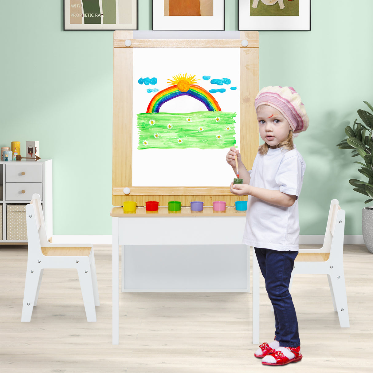 Child standing next to a drawing on an easel with a colorful rainbow and sun.