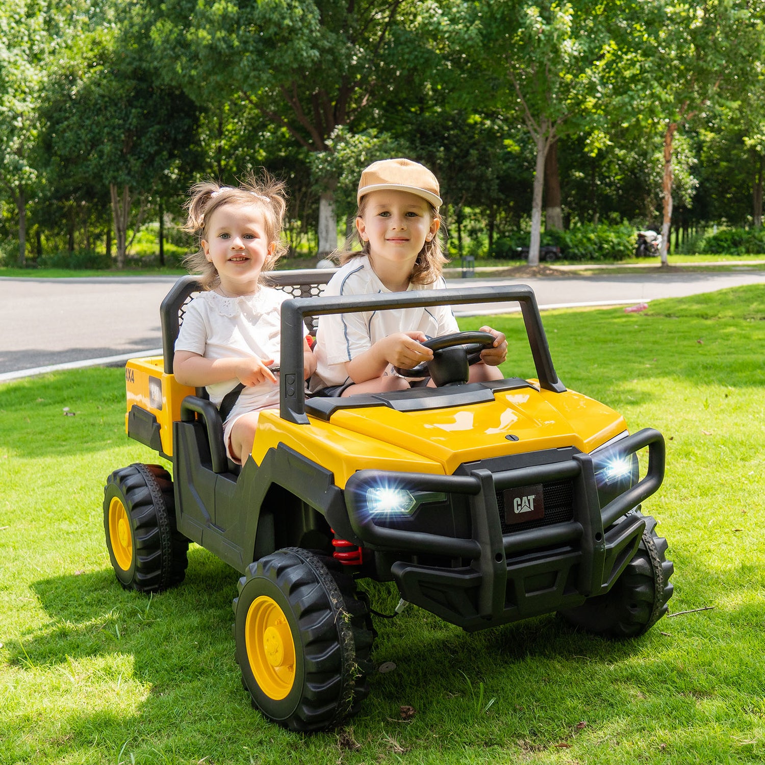 Two children sitting in a yellow and black toy off-road vehicle on grass with trees in the background